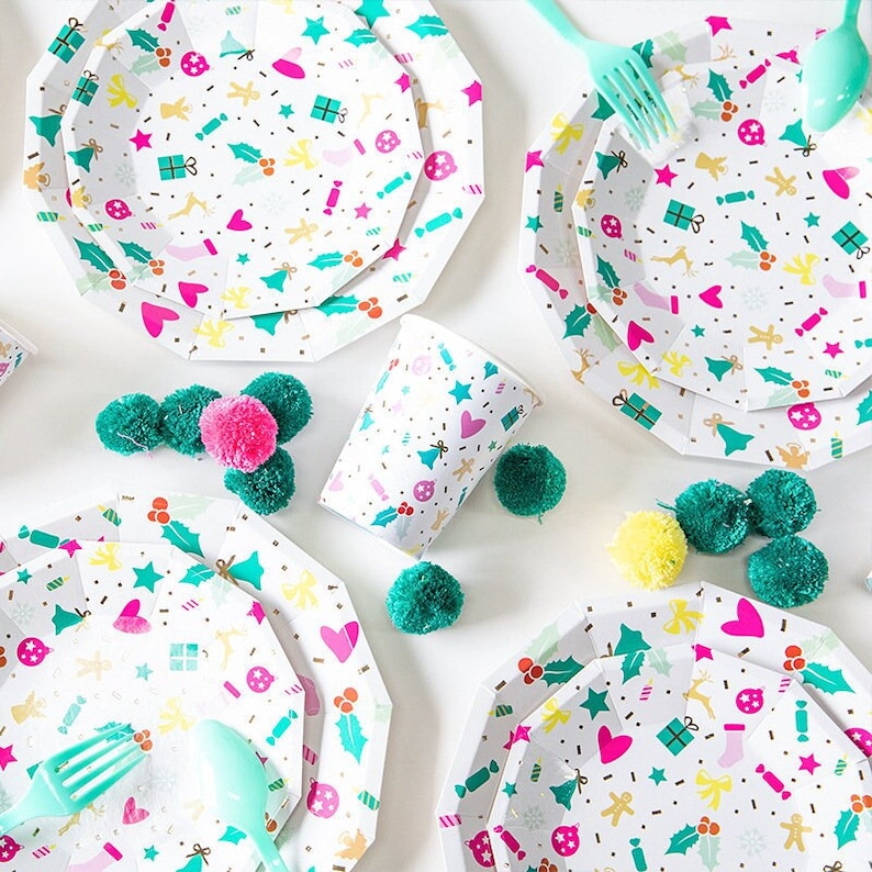 May include: White octagonal paper plates, a cup, and teal plastic utensils are decorated with a festive pattern of hearts, stars, and gift boxes. Green, pink, and yellow pom-poms are scattered around the party supplies.