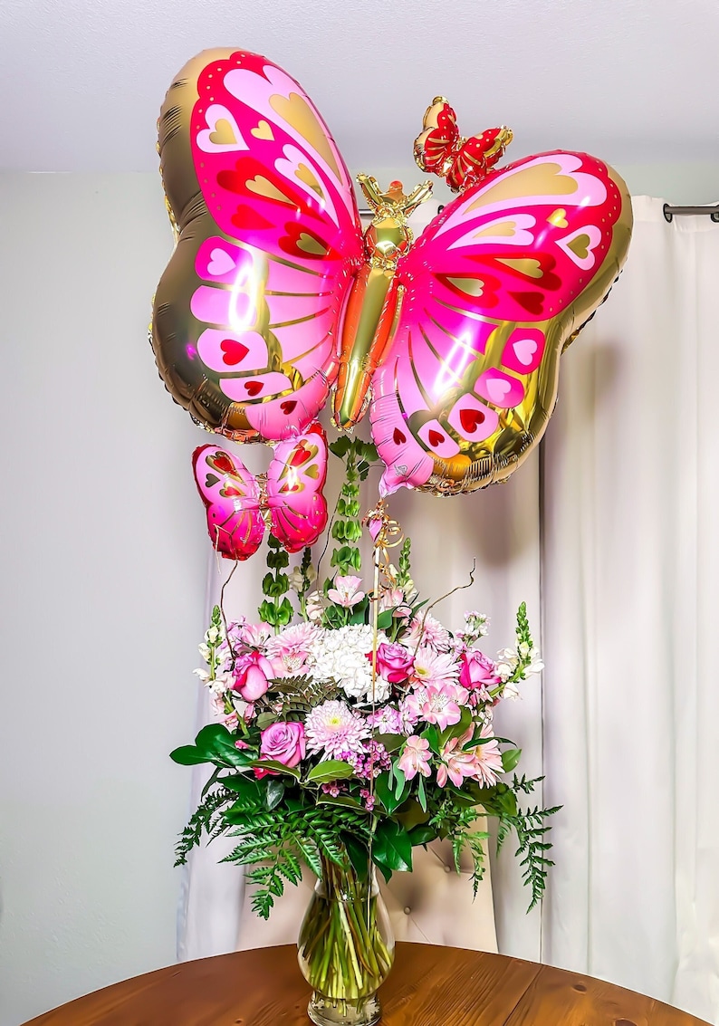 May include: A bouquet of pink and white flowers in a clear glass vase with three pink and gold butterfly balloons attached.