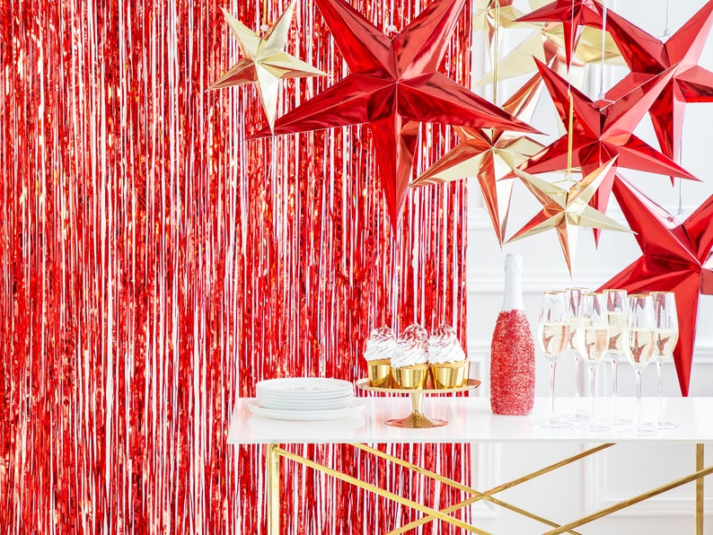 Op de afbeelding: Een tafelsetting met rode en gouden folie sterdecoraties, champagneflutes, een fles mousserende wijn en cupcakes. De tafel is gedekt op een rode folie achtergrond.