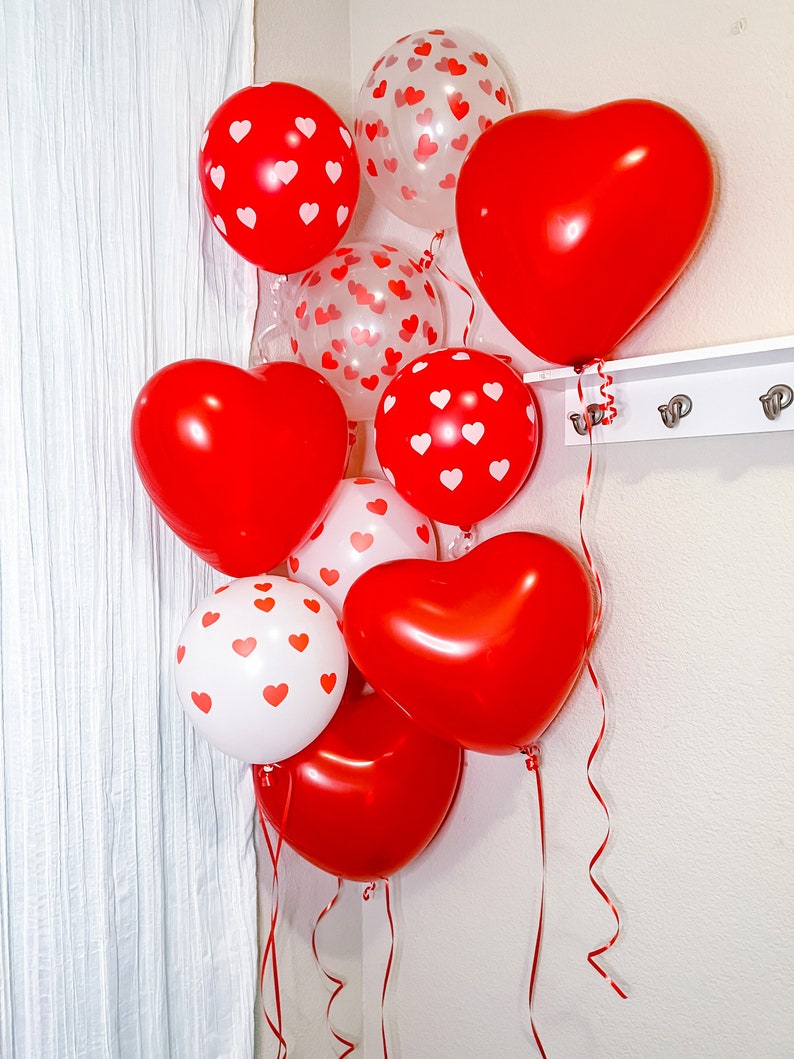 May include: A cluster of red and white heart-shaped balloons with white polka dots. The balloons are tied together with red ribbon and are hanging from a white wall.