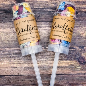 K&ouml;nnte beinhalten: Zwei transparente Konfettikanonen aus Kunststoff, gef&uuml;llt mit getrockneten Bl&uuml;tenbl&auml;ttern. Die Kanonen haben braune Papieretiketten mit der Aufschrift "posh and sparkle Floralfet  Flower Petal Confetti Eco-Friendly Celebration Push-Pop".