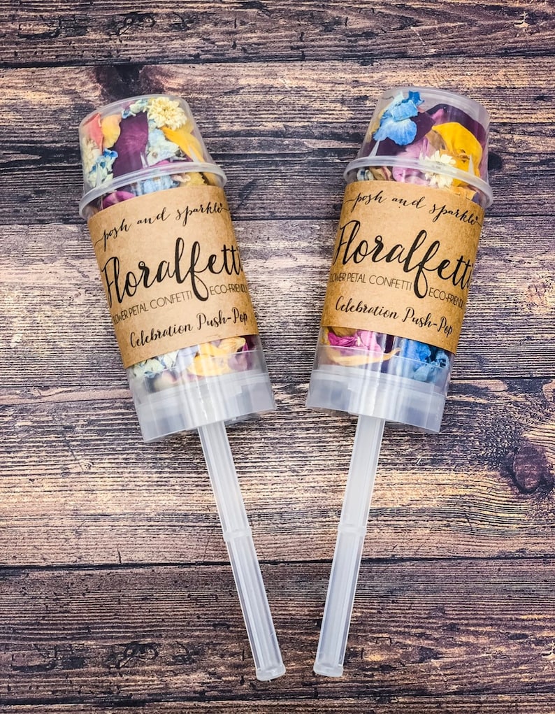 K&ouml;nnte beinhalten: Zwei transparente Konfettikanonen aus Kunststoff, gef&uuml;llt mit getrockneten Bl&uuml;tenbl&auml;ttern. Die Kanonen haben braune Papieretiketten mit der Aufschrift "posh and sparkle Floralfet  Flower Petal Confetti Eco-Friendly Celebration Push-Pop".