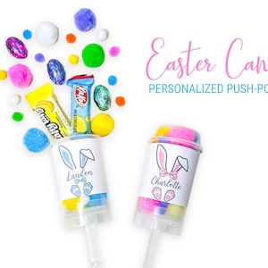 May include: Easter-themed push-pop candy containers with bunny ears and personalized names. The image features colorful candies, pom-poms, and chocolate bars. The text on the image reads "Easter Candy" and "Personalized Push-Pops."