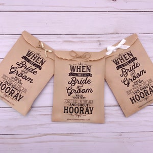 May include: Three brown paper bags with the text "When the Bride and Groom" printed on them. The bags are decorated with twine and a small bow. The text on the bags instructs guests to toss the bags in the air and shout "Hooray!" when the bride and groom walk down the aisle.