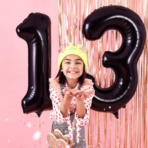 May include: A person celebrates their thirteenth birthday with large black number balloons. They wear a yellow beanie with a crown, a red and white heart-patterned shirt, and a striped skirt. Confetti floats against a pink backdrop with gold streamers.