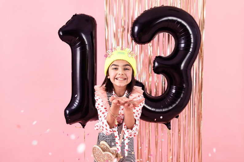 May include: A person celebrates their thirteenth birthday with large black number balloons. They wear a yellow beanie with a crown, a red and white heart-patterned shirt, and a striped skirt. Confetti floats against a pink backdrop with gold streamers.