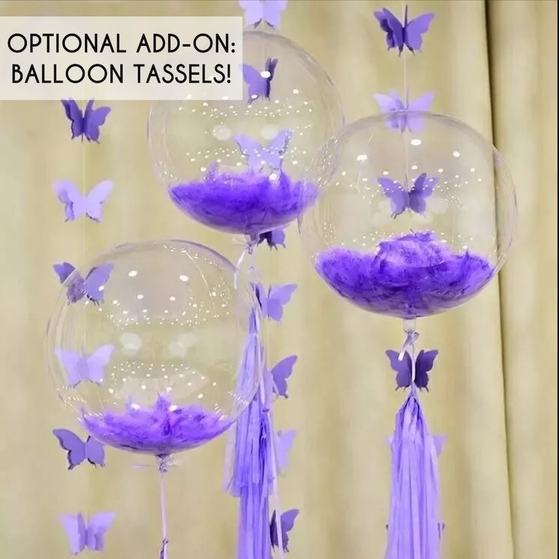 May include: Three clear plastic balloons with purple feathers inside. Each balloon has a string of purple paper butterflies attached to it. The balloons are tied together with purple tassels.