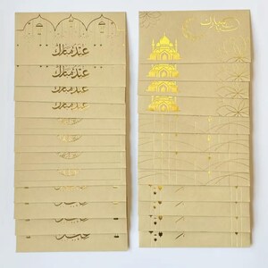 May include: A stack of beige greeting cards with gold foil accents. The cards feature Arabic calligraphy and architectural designs. The cards are for the holiday Eid Mubarak.