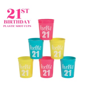 May include: Six plastic shot glasses in yellow, pink, and blue colors. Each glass has the text "hello 21" printed on it. The glasses are stacked in a pyramid shape. 21st Birthday Plastic Shot Cups.