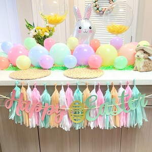 May include: A pastel colored Easter party decoration with a banner that reads "Happy Easter" and a bunny balloon. The banner is made of tissue paper tassels and the letters are cut out of paper. There are also pastel colored balloons and a wreath with pastel colored eggs.