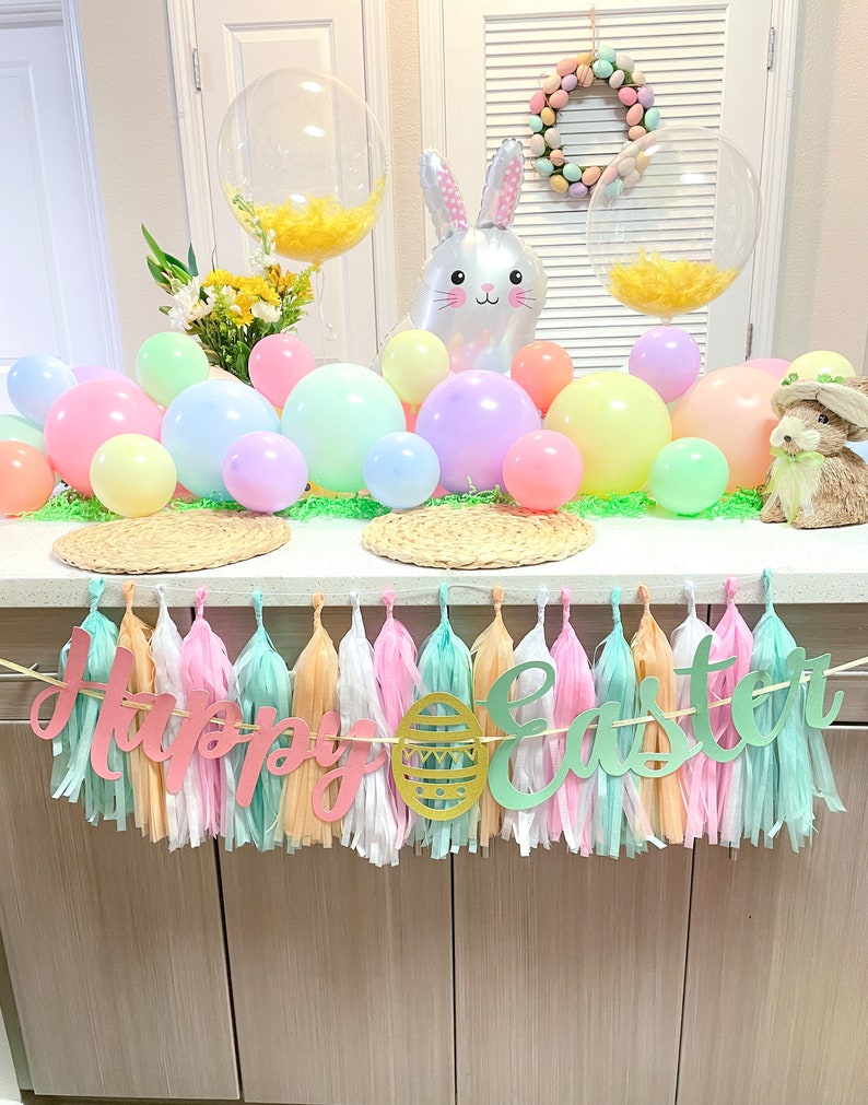 May include: A pastel colored Easter party decoration with a banner that reads "Happy Easter" and a bunny balloon. The banner is made of tissue paper tassels and the letters are cut out of paper. There are also pastel colored balloons and a wreath with pastel colored eggs.