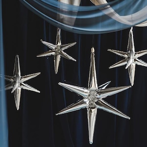 May include: Five silver star-shaped balloons hanging from a blue fabric backdrop. The balloons are inflated and have a metallic sheen.