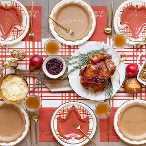 May include: Thanksgiving table setting with a roasted turkey, pumpkin pie, and various side dishes. The table is decorated with leaf-shaped plates, gold cutlery, and a red and white plaid tablecloth. The word "thankful" is written on some plates.
