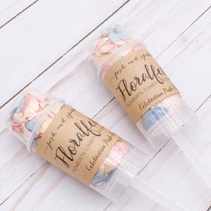 K&ouml;nnte beinhalten: Zwei durchsichtige Plastikr&ouml;hrchen gef&uuml;llt mit pastellrosa, blauen und wei&szlig;en Bl&uuml;tenbl&auml;ttern. Die R&ouml;hrchen haben braune Papieretiketten mit der Aufschrift "posh and sparkly Floralfy Flower Petal Confetti Decorations Celebration Push-Up Confetti".