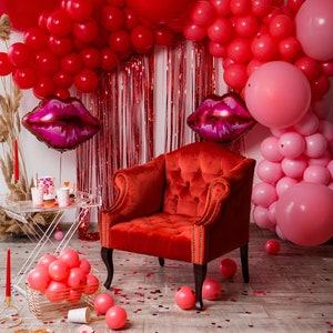 Op de afbeelding: Een rode fluwelen fauteuil staat voor een muur die is versierd met rode en roze ballonnen, rode tinsel en twee grote roze lippenballonnen. De vloer is bedekt met rode confetti.
