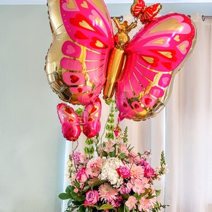 May include: A bouquet of pink and white flowers in a clear glass vase with three pink and gold butterfly balloons.