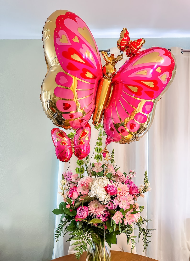 May include: A bouquet of pink and white flowers in a clear glass vase with three pink and gold butterfly balloons.