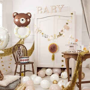 Pu&ograve; includere: Una decorazione per una festa di baby shower con un palloncino a forma di orso marrone, un palloncino bianco con la scritta "Oh Baby", frange dorate in lamina, un cartello in legno con la scritta "BABY", un armadio bianco, un tavolo con un porta torta e un ventaglio di carta rosa e oro.