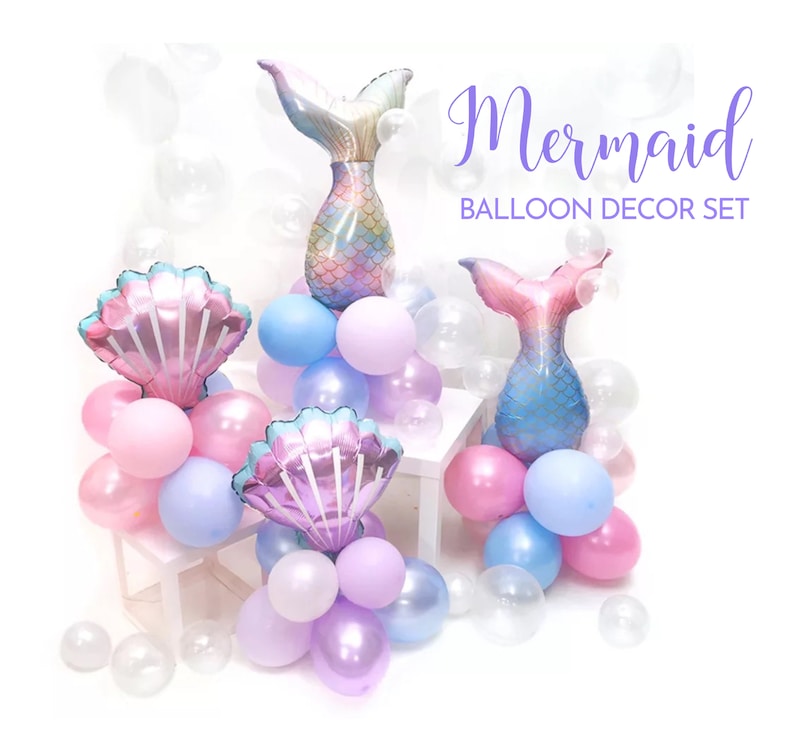 May include: A mermaid-themed balloon decor set with two foil mermaid tail balloons, two foil seashell balloons, and a variety of pastel-colored latex balloons. The balloons are arranged on white stands and include clear balloons.