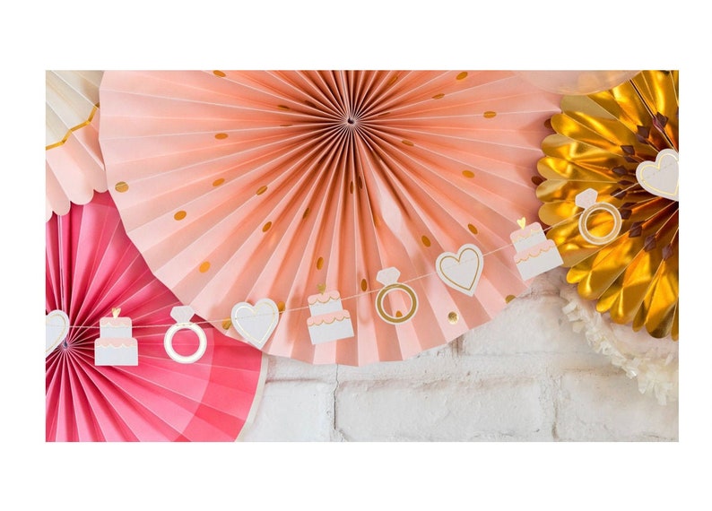 K&ouml;nnte beinhalten: Eine wei&szlig;e und goldene Papiergirlande mit Herzen, Ringen und Kuchenmotiven. Die Girlande h&auml;ngt an einer wei&szlig;en Backsteinwand mit rosa und goldenen Papierf&auml;chern dahinter.