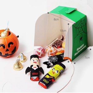 May include: An orange plastic jack-o'-lantern bucket filled with gold-wrapped candies, alongside a green cardboard box labeled Happy Halloween with dimensions 8cm by 10cm by 7cm, containing assorted sweets, a vampire figure, and a monster figure.