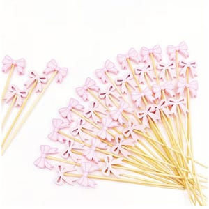 May include: A collection of light pink bow-shaped decorative picks on wooden skewers. The bows are a solid pastel pink color, and the skewers are a natural wood tone. These are likely for use in cupcakes or other desserts.