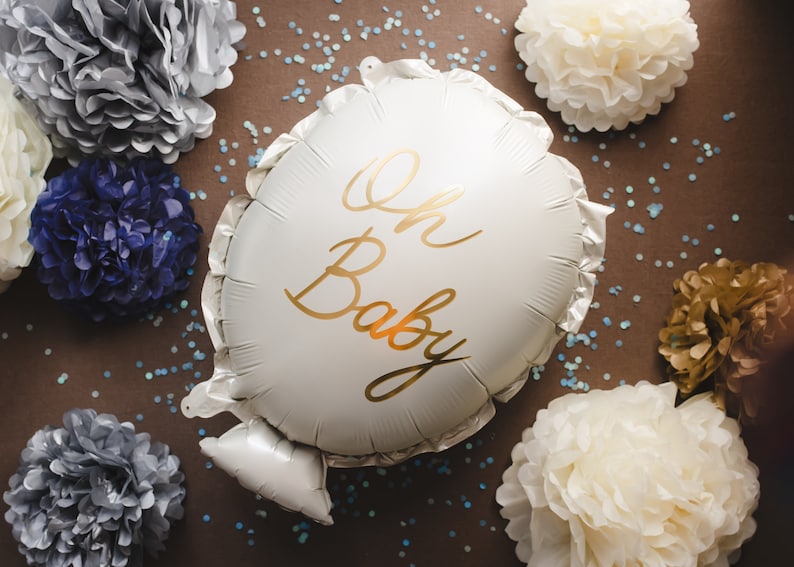 May include: A white balloon with the text "Oh Baby" written in gold script. The balloon is surrounded by white, gold, and blue tissue paper pom-poms.