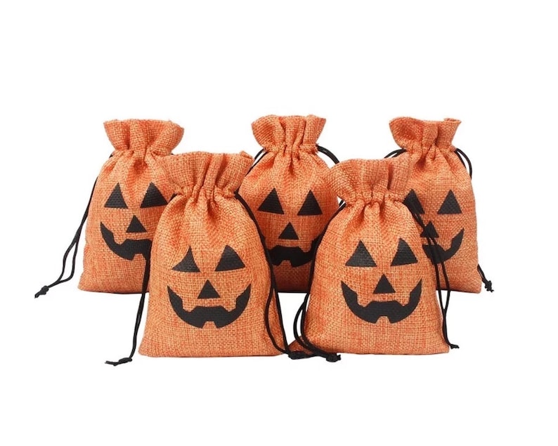 May include: A set of five orange burlap drawstring bags, each featuring a black printed jack-o'-lantern face. The bags have a textured fabric weave and are cinched at the top with thin black cords, creating a gathered opening for each pouch.