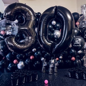 May include: A black and silver 30th birthday party decoration set. The set includes large black number balloons, smaller black, silver, and pink balloons, and balloons printed with "RIP Youth". There are also black cups and bottles.