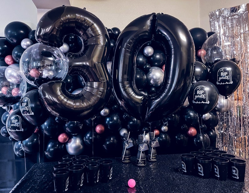 May include: A black and silver 30th birthday party decoration set. The set includes large black number balloons, smaller black, silver, and pink balloons, and balloons printed with "RIP Youth". There are also black cups and bottles.