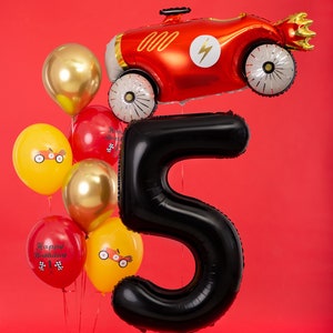 May include: A celebratory balloon display for a fifth birthday. It includes a large black number 5 balloon, a red race car balloon, and a cluster of smaller balloons in gold, red, and yellow. Some balloons have race car designs and "Happy Birthday" text.