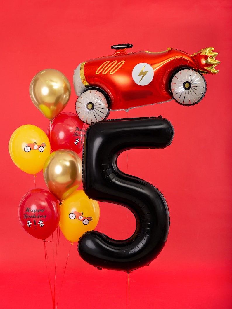 May include: A celebratory balloon display for a fifth birthday. It includes a large black number 5 balloon, a red race car balloon, and a cluster of smaller balloons in gold, red, and yellow. Some balloons have race car designs and "Happy Birthday" text.