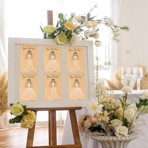 May include: A white framed sign on a wooden easel displaying six rectangular cards with the text Guess The Dress and a black line drawing of a bride in a gown. The sign is decorated with artificial cream and pale green roses and foliage.