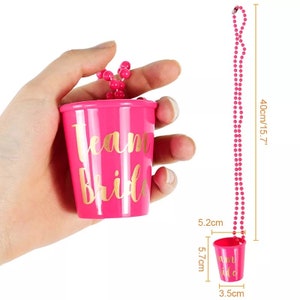 Pu&ograve; includere: Un bicchierino di plastica rosa con l'iscrizione dorata "Team Bride", attaccato a una collana di perline rosa. La collana &egrave; lunga 40 cm. Il bicchierino &egrave; largo 5,2 cm e alto 5,7 cm.
