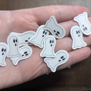May include: A collection of white ghost stickers with black eyes and mouths. The stickers are scattered on a hand.