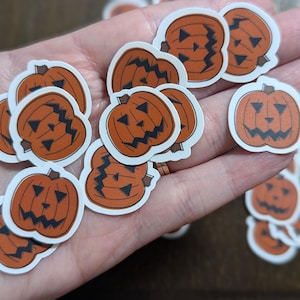 May include: A collection of orange pumpkin stickers with black facial features and white borders. The stickers are in various sizes and are scattered across a surface, perfect for Halloween crafts and decorations.