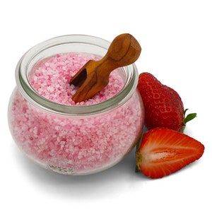 Op de afbeelding: Een glazen pot gevuld met roze aardbeienzout, een klein houten schepje en twee verse aardbeien.