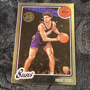 May include: A basketball trading card featuring Koby Brea of the Phoenix Suns. The card has a gold border and includes the Suns logo, the Topps logo, and the text "RC". The card commemorates the 45th anniversary.