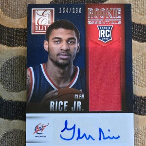 May include: A Glen Rice Jr. rookie card from the 2013-2014 season. The card features a portrait of the basketball player, a red fabric swatch, and his signature. The card is numbered 154/299.