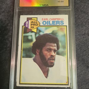 May include: A 1979 Topps trading card of Earl Campbell, graded VG-EX, encased in a protective holder. The card features a portrait of Campbell in his Oilers uniform, with the team name and "All Pro" designation.