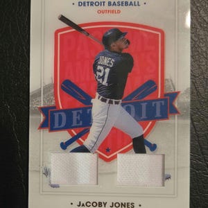 May include: A baseball card featuring Jacoby Jones, an outfielder for the Detroit Tigers. The card has a red, white, and blue design with the team logo and Jones's name and number. The card also includes two patches of white fabric.