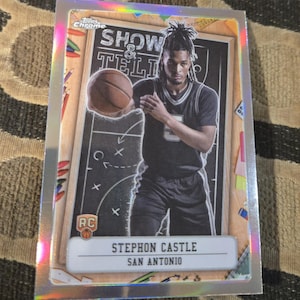 May include: A basketball trading card featuring Stephon Castle of the San Antonio Spurs. The card has a silver border and includes the text "SHOW & TELL" and "RC". The player is depicted holding a basketball in front of a chalkboard.