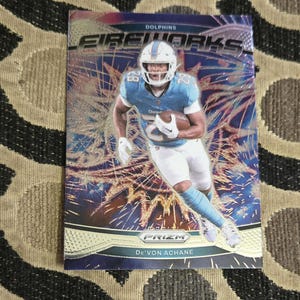 May include: A De'Von Achane football card from the Dolphins team. The card features a player in a blue and white uniform running with the ball. The card has the word "Fireworks" and "Prizm" printed on it.