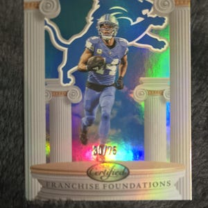 May include: A football trading card featuring a player in a blue uniform, holding a football, with the Detroit Lions logo in the background. The card has a holographic effect and includes the text "30/75" and "Certified Franchise Foundations."