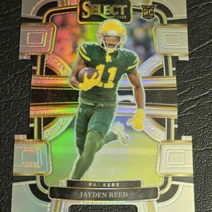 May include: A green and gold football card featuring a player in a green uniform with the number 11. The card has a silver background with a black and gold border. The text "SELECT 2023" is in the upper left corner. The text "PACKERS" and "JAYDEN REED" is in the lower center of the card.