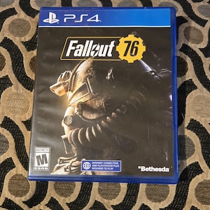 May include: A PS4 game case for Fallout 76. The cover features a detailed illustration of a character in a helmet. The game title is in yellow and white. The case is blue with the ESRB rating visible.