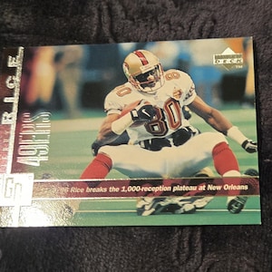 May include: A collectible trading card featuring a football player in a white and red uniform with the number 80. The card has the text "49ERS" and "RICE" on the side. The text on the card reads "96 Rice breaks the 1,000-reception plateau at New Orleans."
