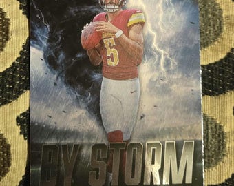Puede incluir: Una tarjeta de intercambio de fútbol americano con un jugador de fútbol en un uniforme rojo y blanco con el número 5 en la parte delantera. La tarjeta tiene un fondo negro con texto metálico plateado que dice "By Storm" y "Jayden Daniels - Commanders". La tarjeta también tiene un logotipo de Rookie Card en la esquina superior derecha.