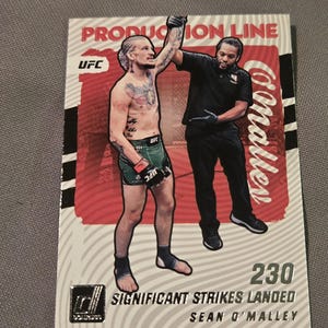 May include: A trading card featuring a UFC fighter with his arm raised in victory. The card has a red and white background with the text "PRODUCTION LINE" and "SIGNIFICANT STRIKES LANDED". The card also includes the number "230".