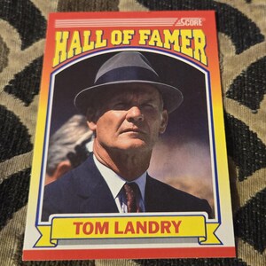 May include: A vintage Tom Landry Hall of Famer trading card. The card features a portrait of Tom Landry wearing a fedora and suit. The card has a red border with the words "HALL OF FAMER" and "TOM LANDRY".